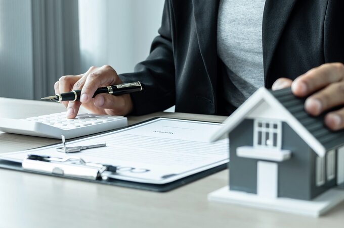 Eine Person sitzt an einem Tisch, arbeitet mit einem Taschenrechner und hält einen Stift. Vor ihr liegt ein Dokument und ein Modell eines Hauses. Die Szene vermittelt ein Gefühl von Immobilienbewertung oder -beratung.